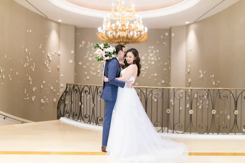 Bride and groom embrace on a marble landing beneath a gold chandelier, curved iron railing and petal-dotted wall behind