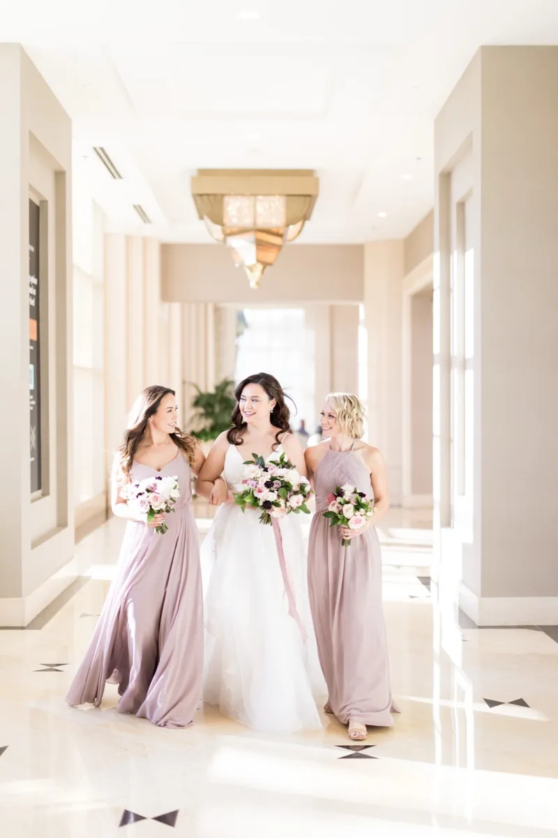 Bride walking a bright hallway with two bridesmaids in mauve, all three holding bouquets
