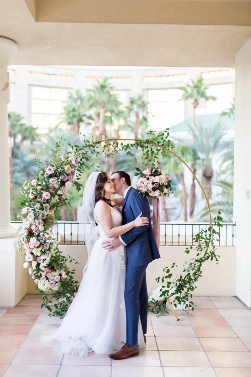 Bride and groom kissing in front of a circular floral arch with daylight and palms behind them