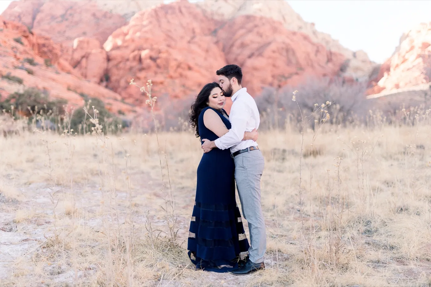 Woman in a long navy dress and her fiancé in a white shirt holding each other in golden grass below the red cliffs of Red Rock Canyon