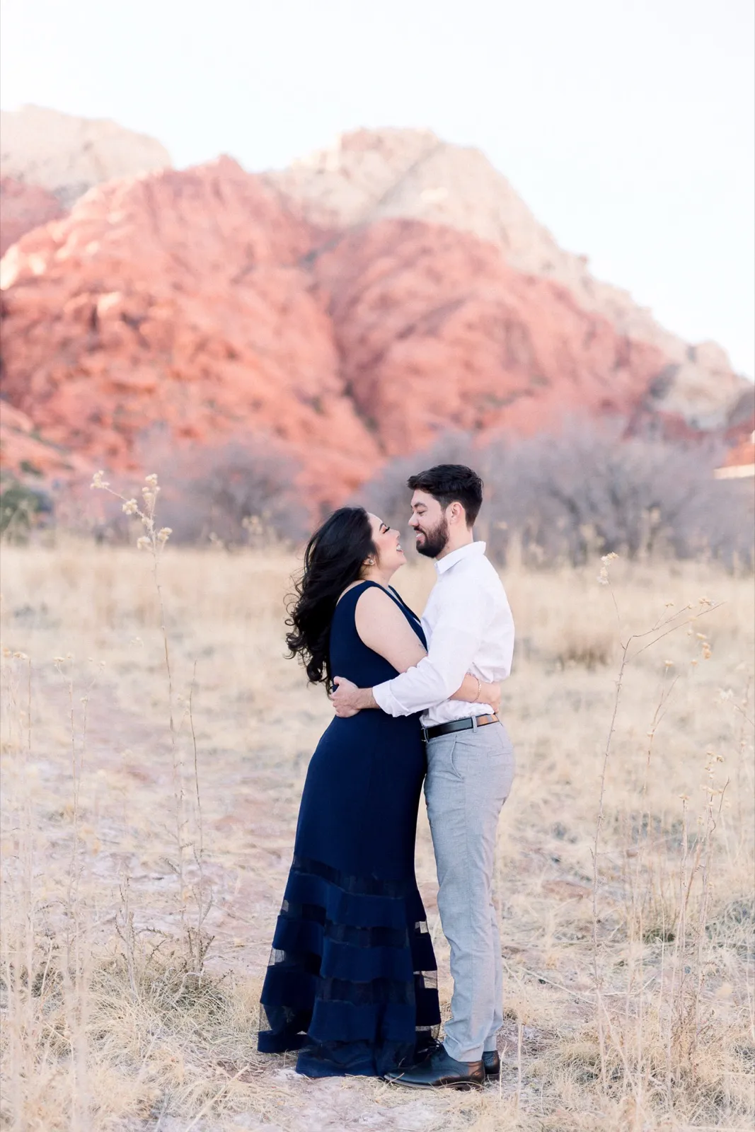 Couple standing close in dry golden grass with the sunlit red and cream cliffs of Red Rock Canyon behind them