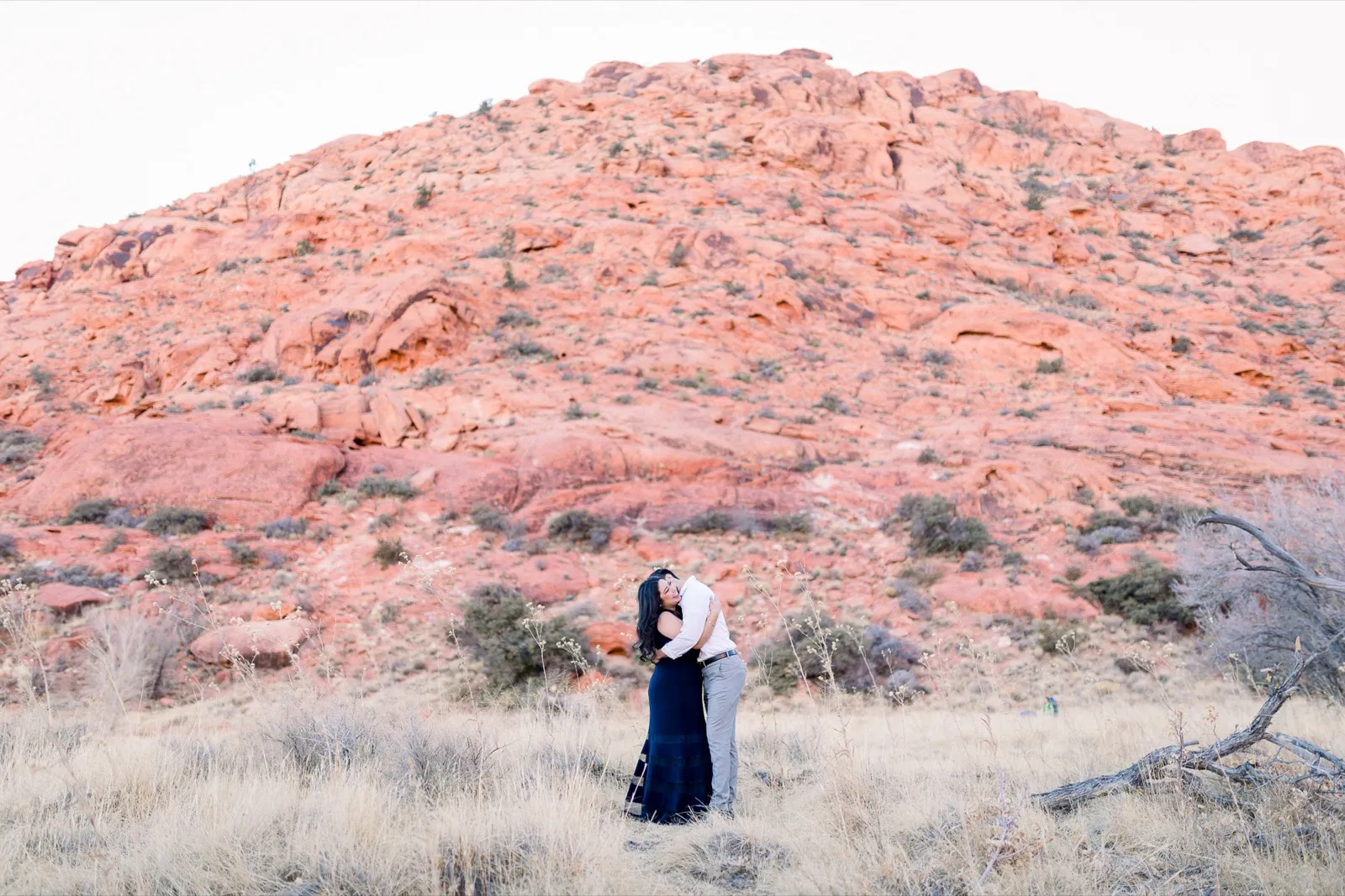 Couple embracing in dry grass, small in the frame below a huge sunlit dome of red sandstone