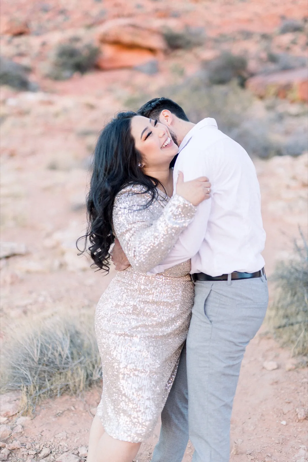 Woman in a champagne sequin dress leaning into her fiancé in a white shirt with red sandstone behind them