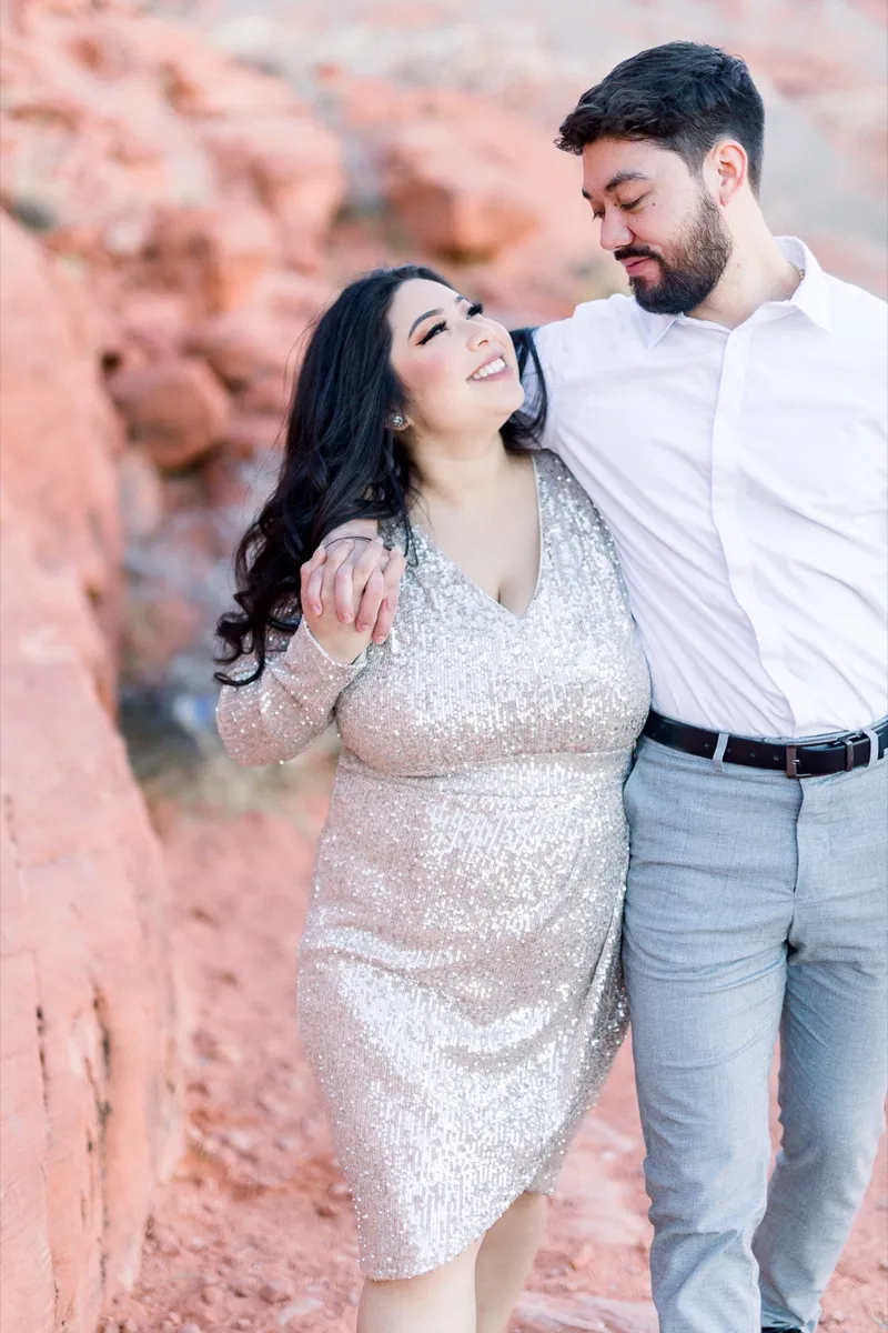 Couple standing side by side against a wall of red rock with her arm across his chest