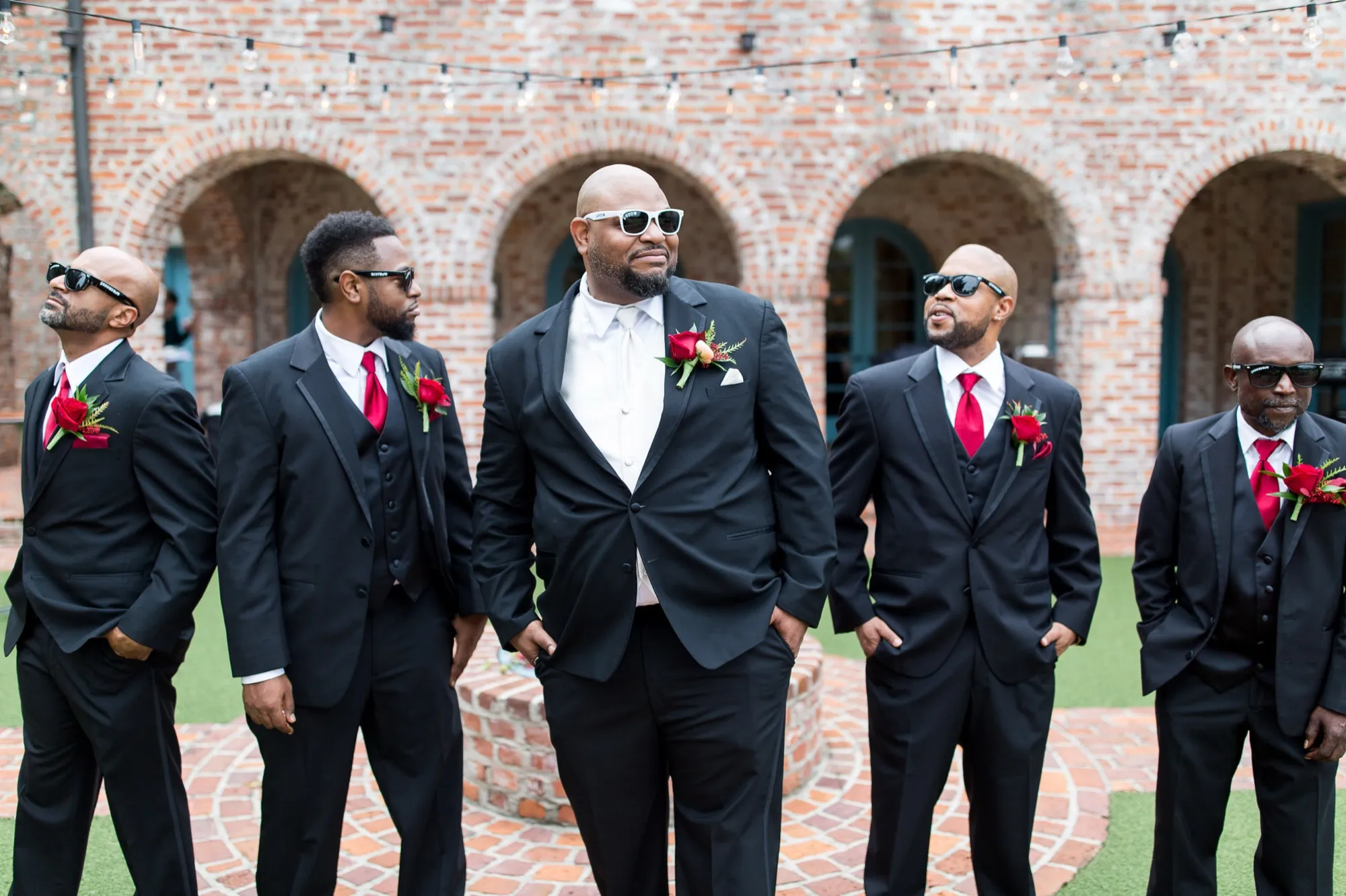 Groom and his groomsmen in black tuxedos with red rose boutonnieres in a brick courtyard