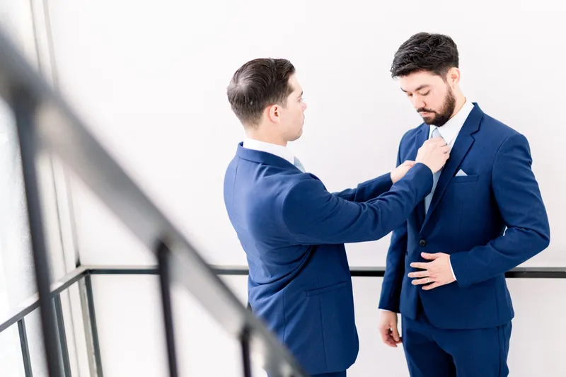 A groomsman in a navy suit straightening the groom's tie against a bright white wall beside a stair railing
