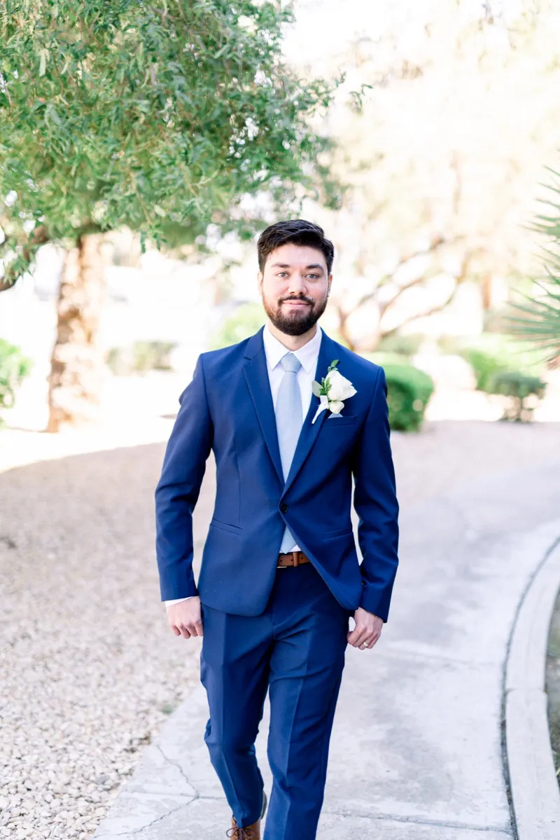 The groom walking along a gravel path outside in a royal blue suit and pale blue tie, olive trees behind him