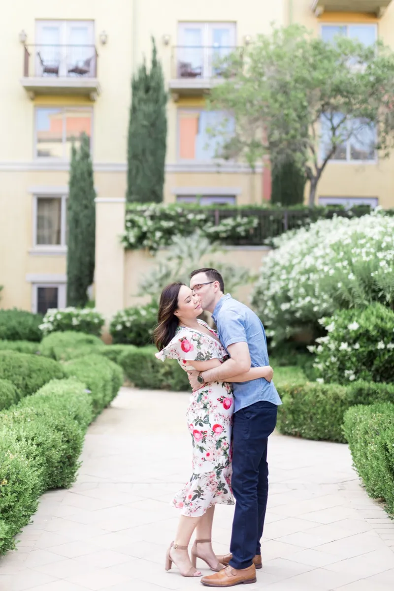 Man in a blue shirt leaning in to kiss his fiancee on a garden path lined with clipped hedges and cypress trees