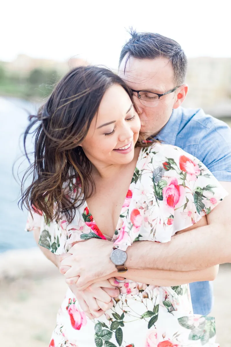 Close portrait of a man holding his fiancee from behind and kissing her temple with the lake blurred behind them