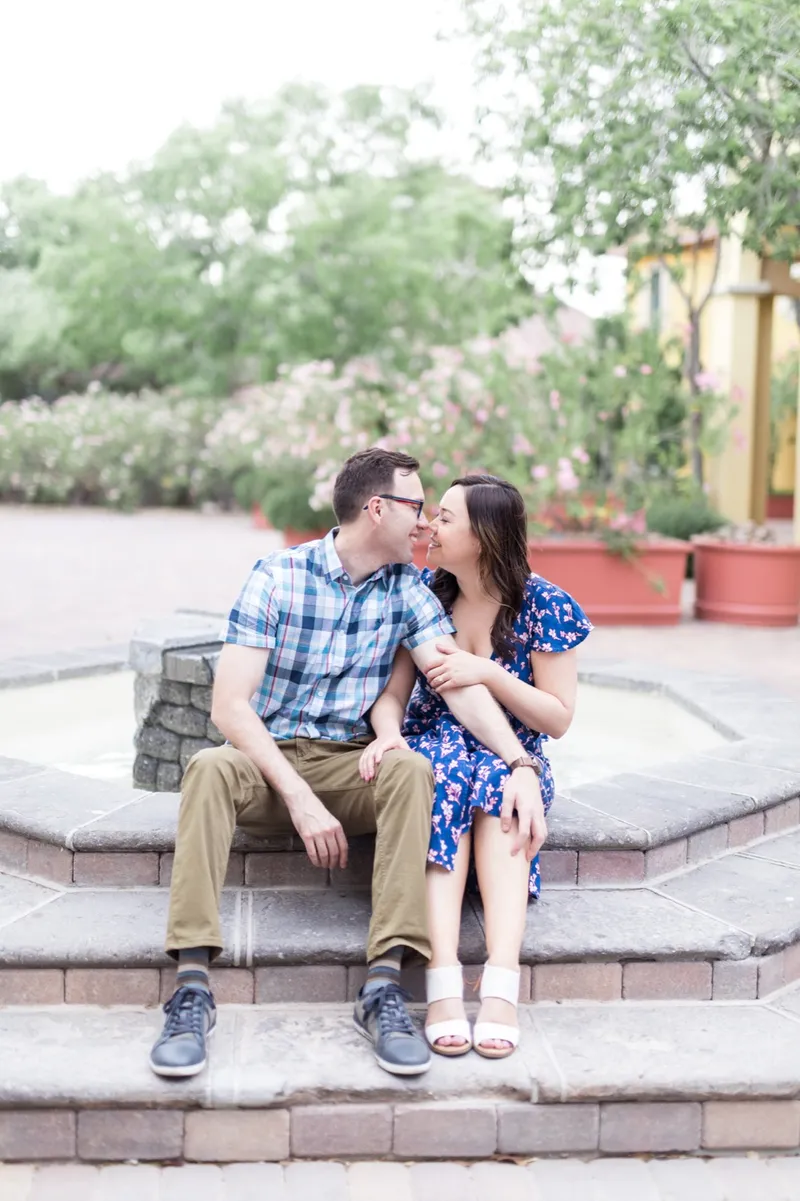 Couple sitting on the steps of a stone fountain leaning in toward each other, him in a blue plaid shirt and her in a blue floral dress