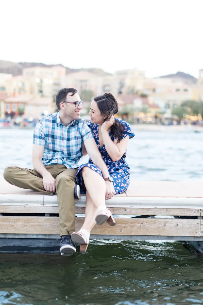 Couple sitting on the edge of a dock with their feet hanging over the water and the village rooftops across the lake