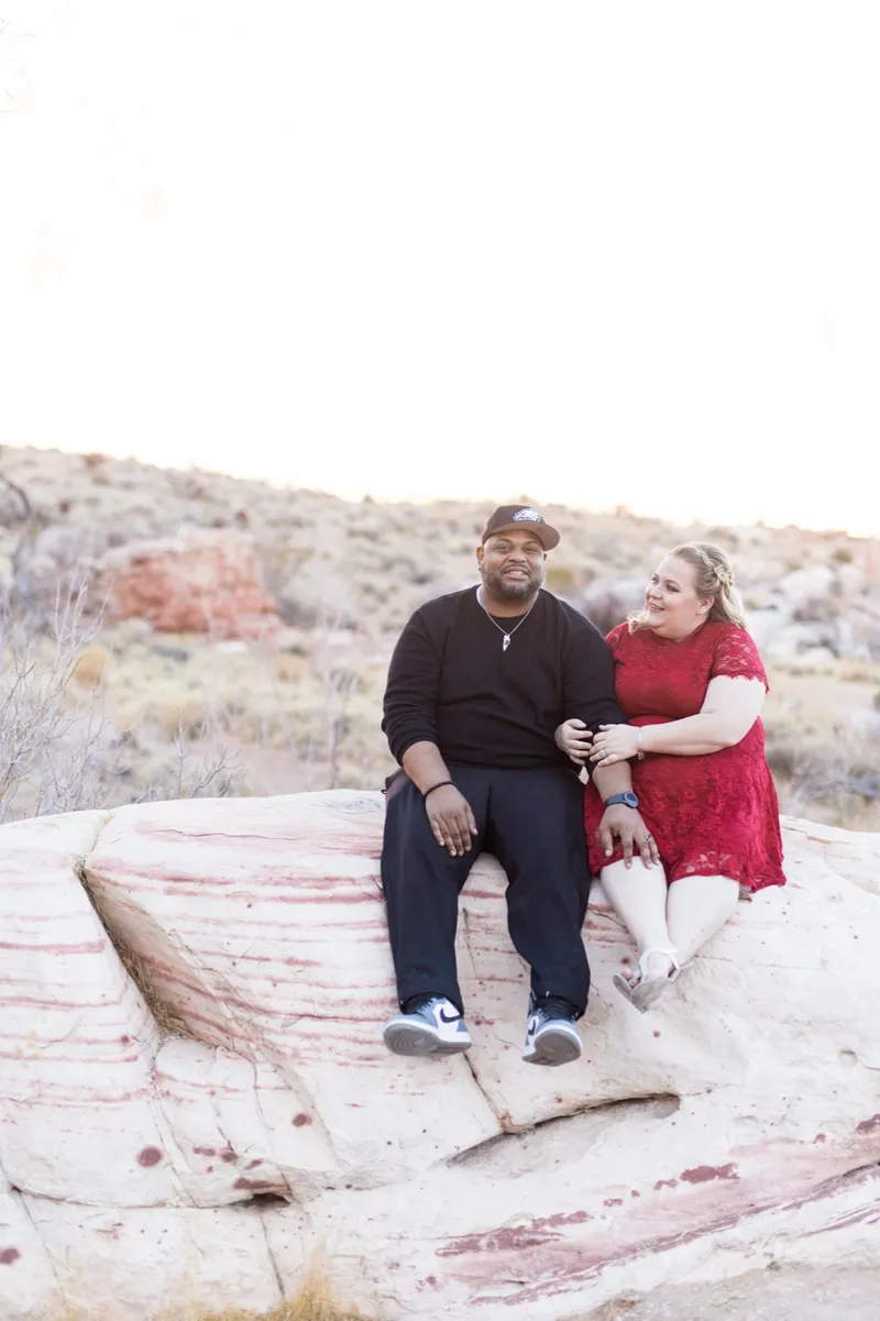 Couple holding hands on a pale sandstone ledge, him seated in dark clothes and her standing in a red lace dress