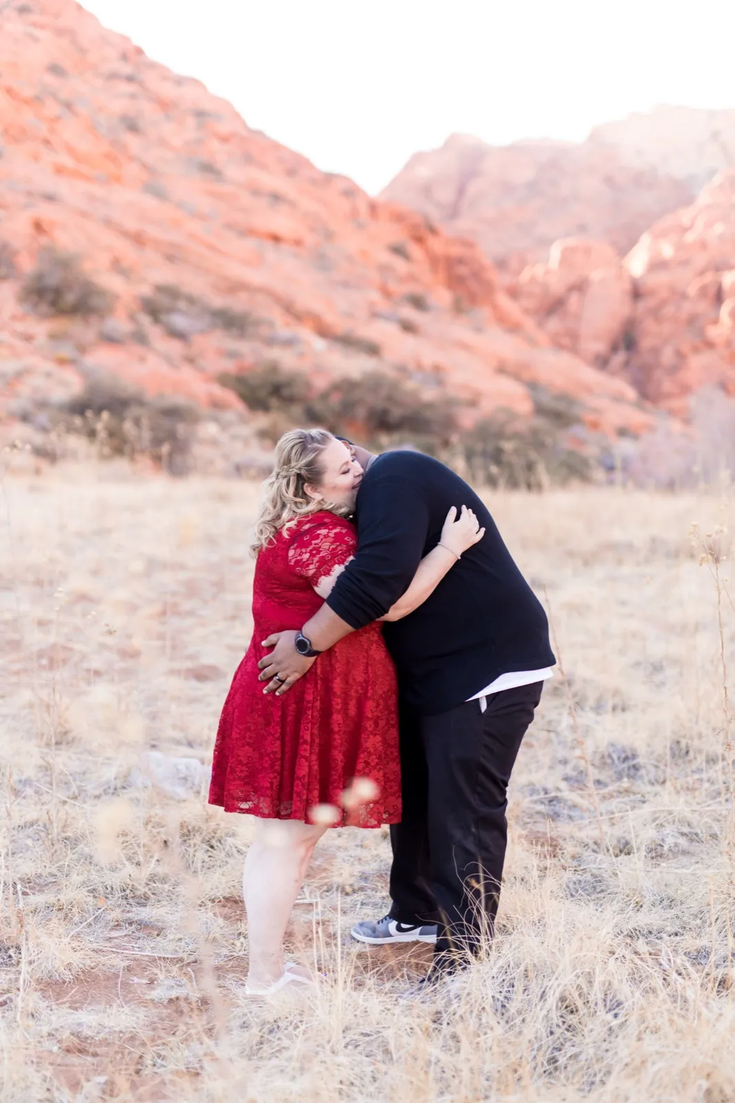Man leaning in to kiss his partner in dry golden grass below the sunlit red Calico cliffs, her in a red lace dress