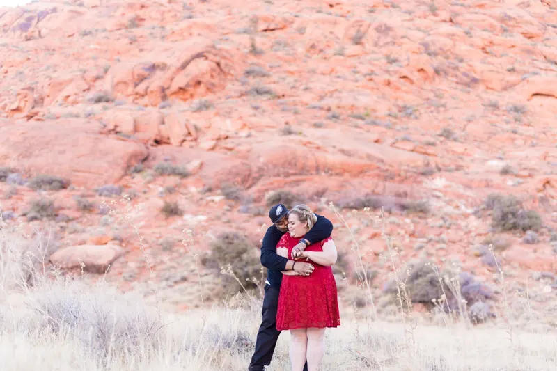 A couple embraces in front of red sandstone cliffs at Red Rock Canyon, her red lace dress bright against the pale grass