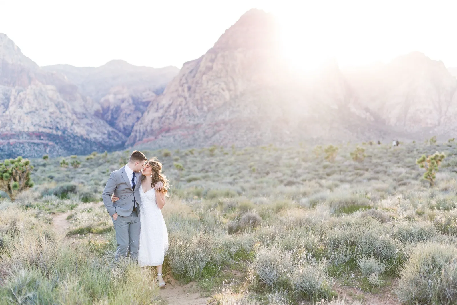 Bride and groom leaning into each other on a sandy path as low sun flares over the peaks behind them