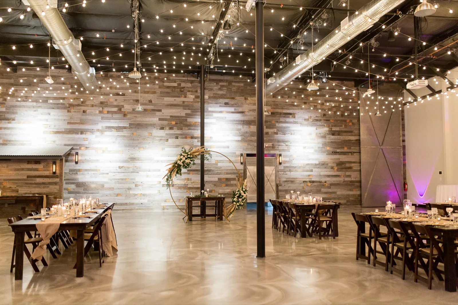 The reception room before anyone walked in, reclaimed wood walls washed with light, a canopy of string lights overhead and a gold hoop arch of white roses and pampas behind the sweetheart table