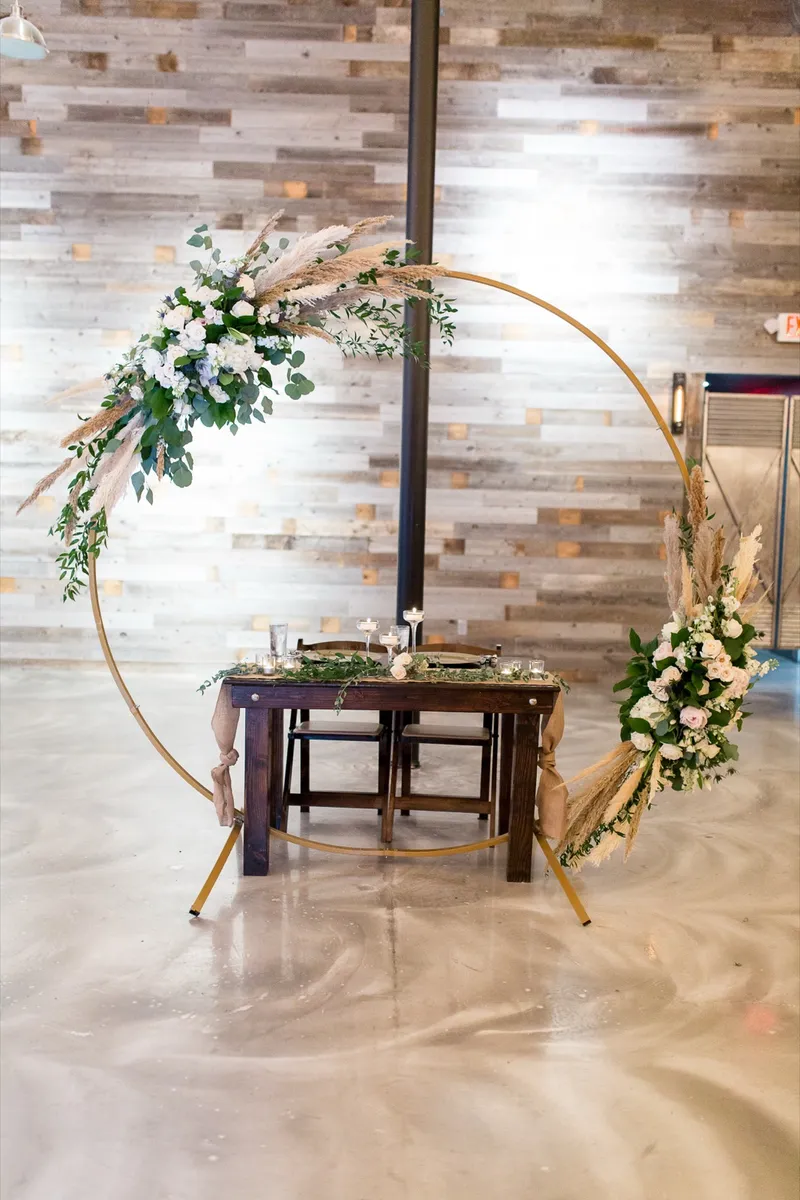 Sweetheart table set inside a tall gold hoop arch wrapped with white roses, eucalyptus and pampas grass against the wood wall
