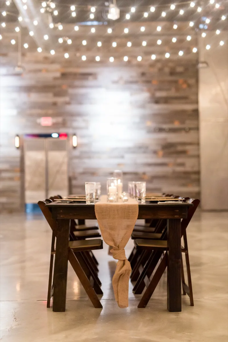 Long farm table set with a burlap runner, candles in glass and dark wood folding chairs on the polished concrete floor