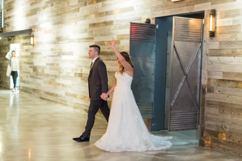 Bride raising her hand and waving as the groom leads her past the metal barn door into the reception