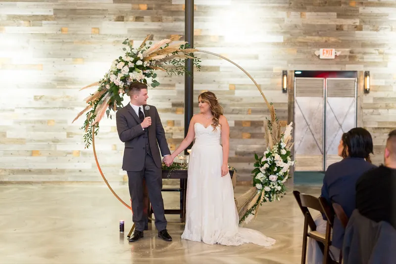 Groom holding a microphone beside the bride in front of the hoop arch while guests listen from the tables