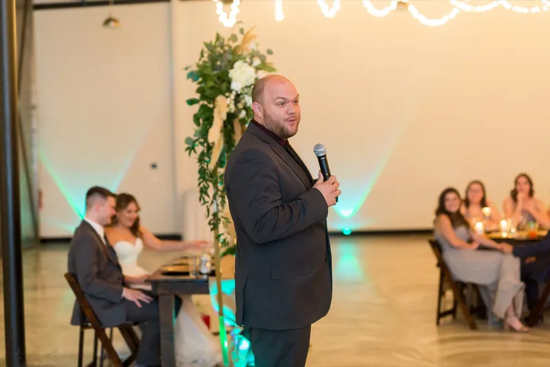 A guest speaking into the microphone during a toast while the couple listen from their table under teal uplight
