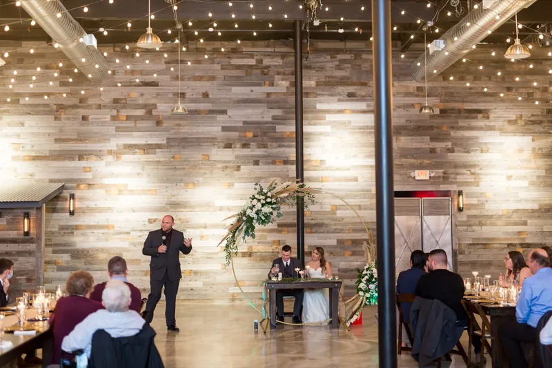 Wide view of the whole room during a toast, the couple seated inside the hoop arch and guests at candlelit tables down both sides
