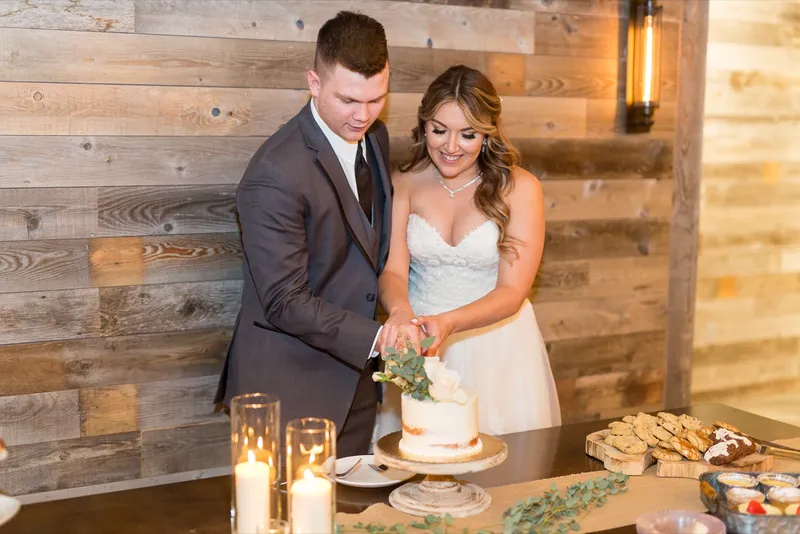 Bride and groom cutting a small naked cake on a wood stand beside rows of candles and trays of desserts