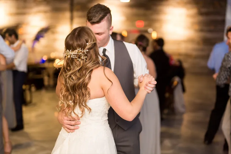 The couple slow dancing among their guests late in the night with the wood wall going soft behind them