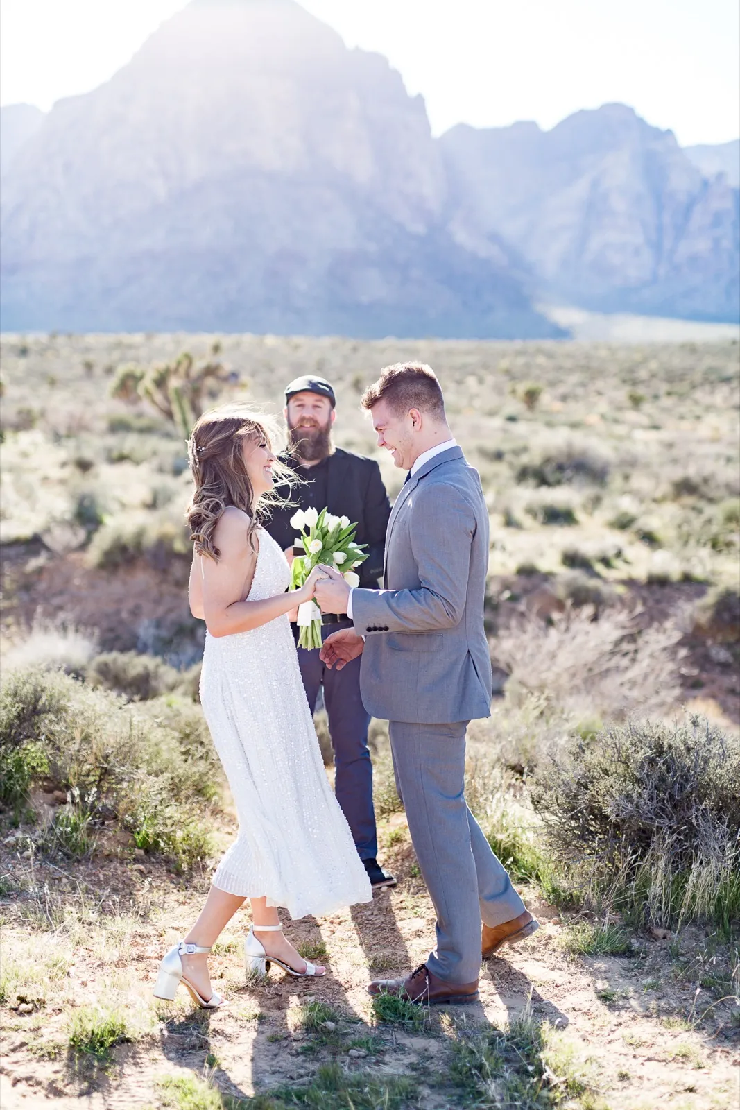 Bride with white tulips and groom in grey stand with their officiant in the brush below the Red Rock cliffs