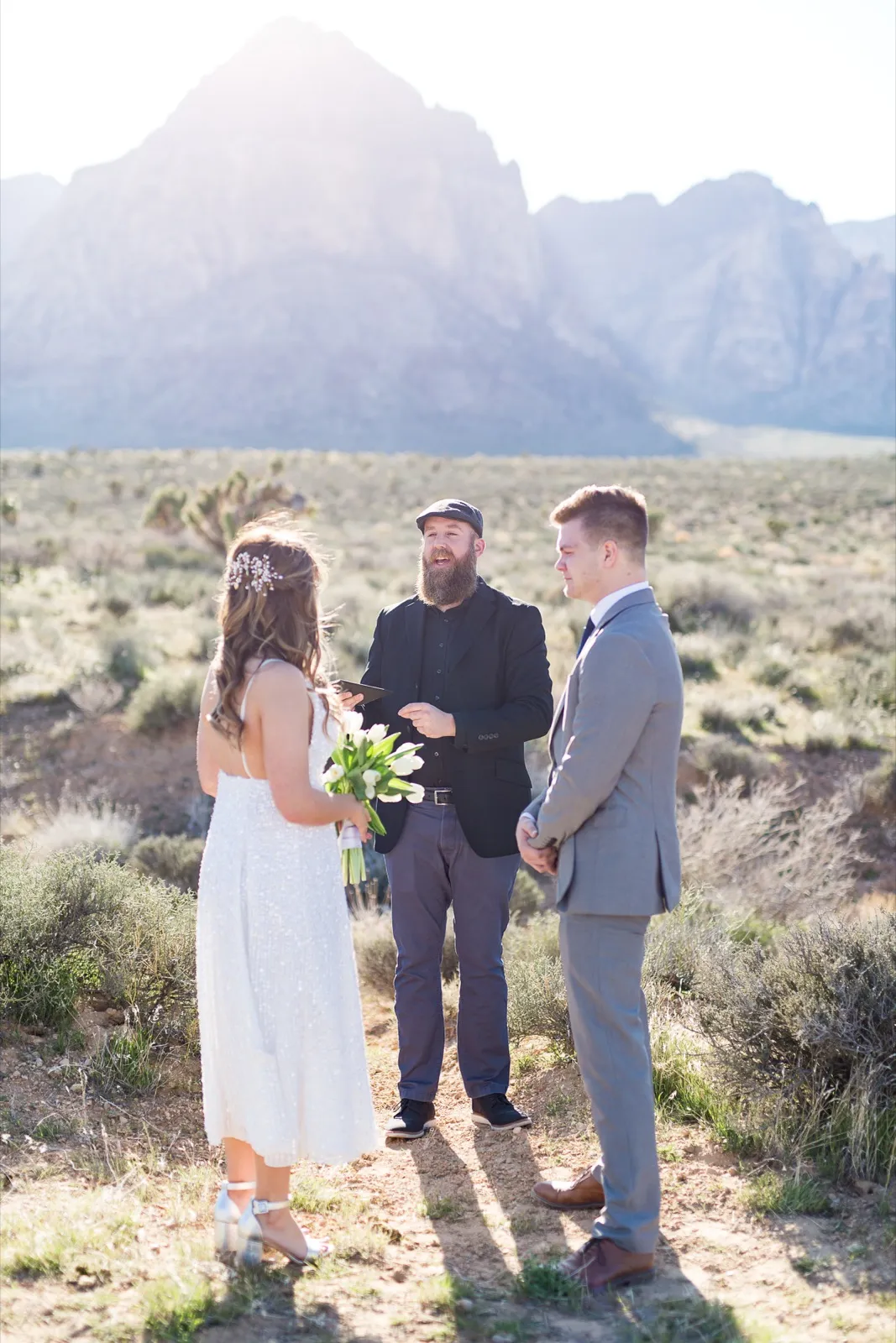 A bride with white tulips and her groom stand with their officiant in open sagebrush below the Red Rock escarpment