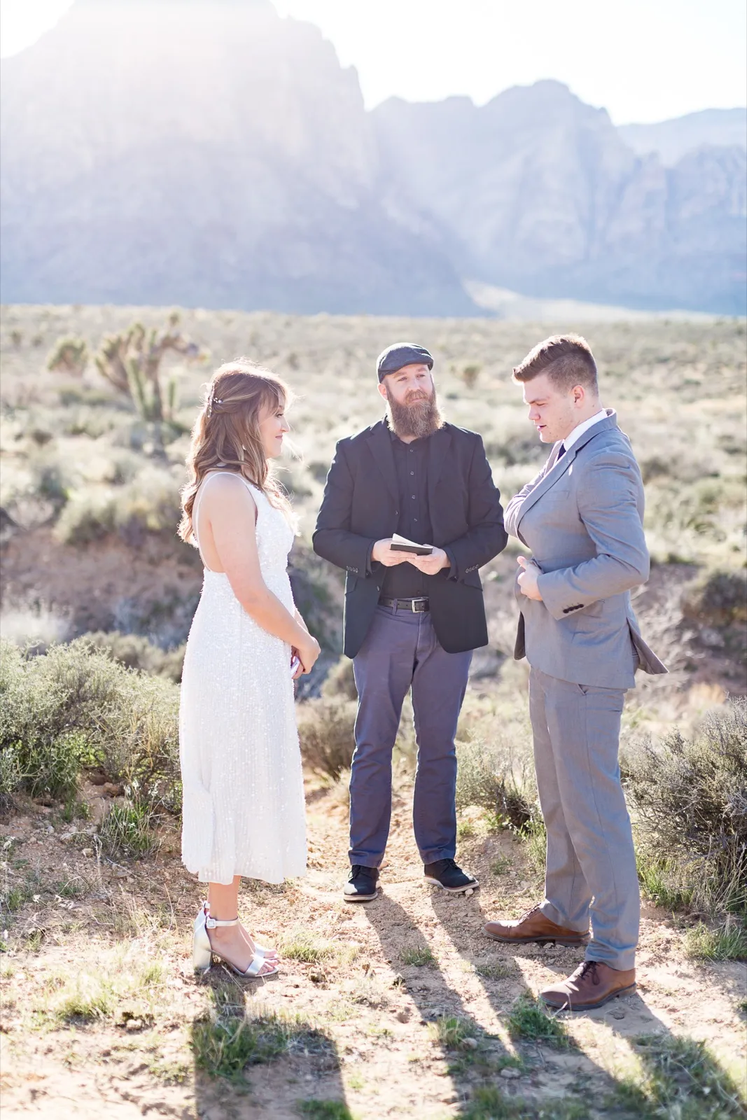 Officiant in a dark jacket and hat reads to a bride and groom standing alone together in the desert brush