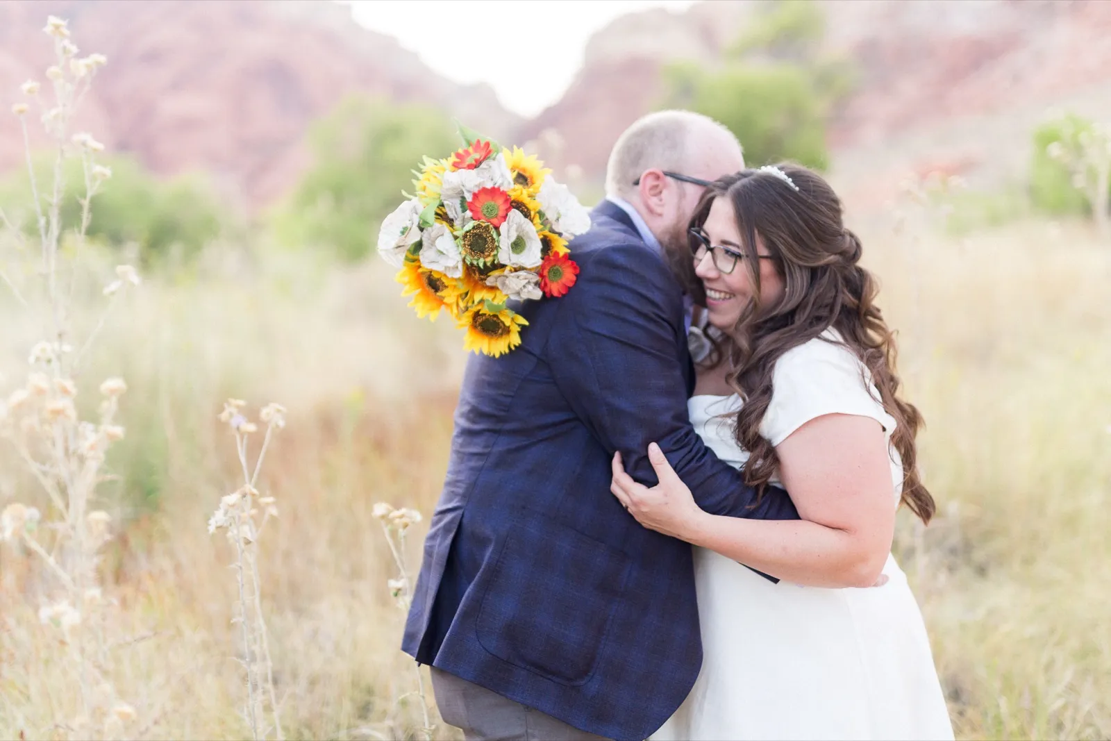 Groom in a navy plaid jacket hugging his bride as she holds a big sunflower bouquet in dry desert grass