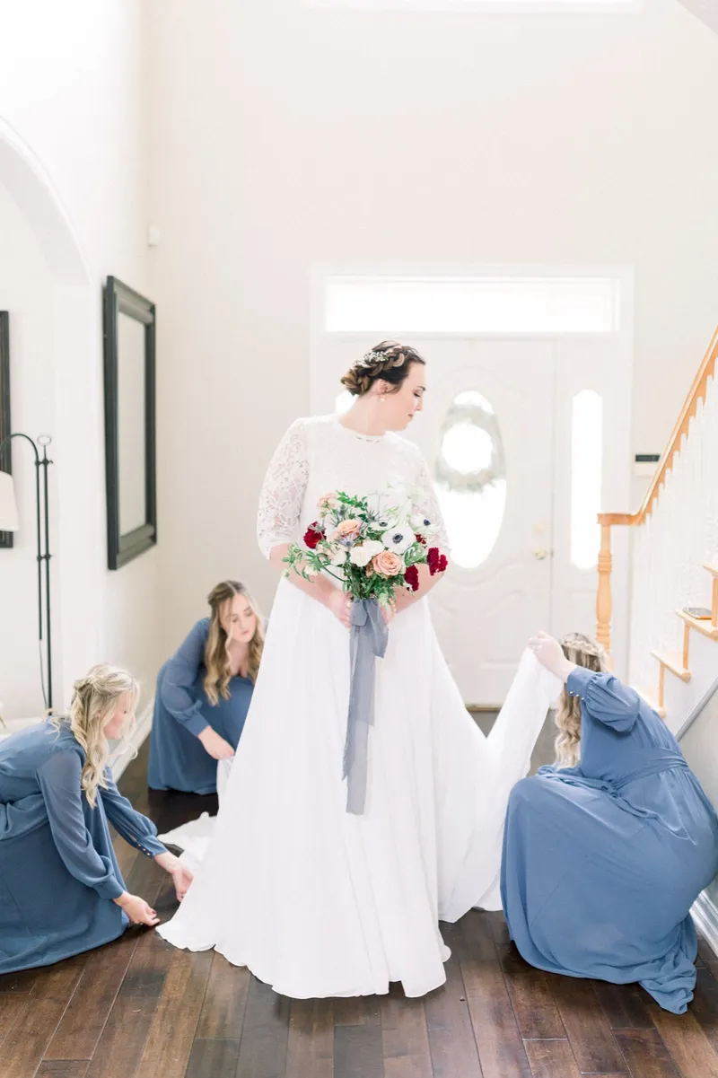 Two bridesmaids in blue crouched on the wood floor of the entryway straightening the train of the bride's gown