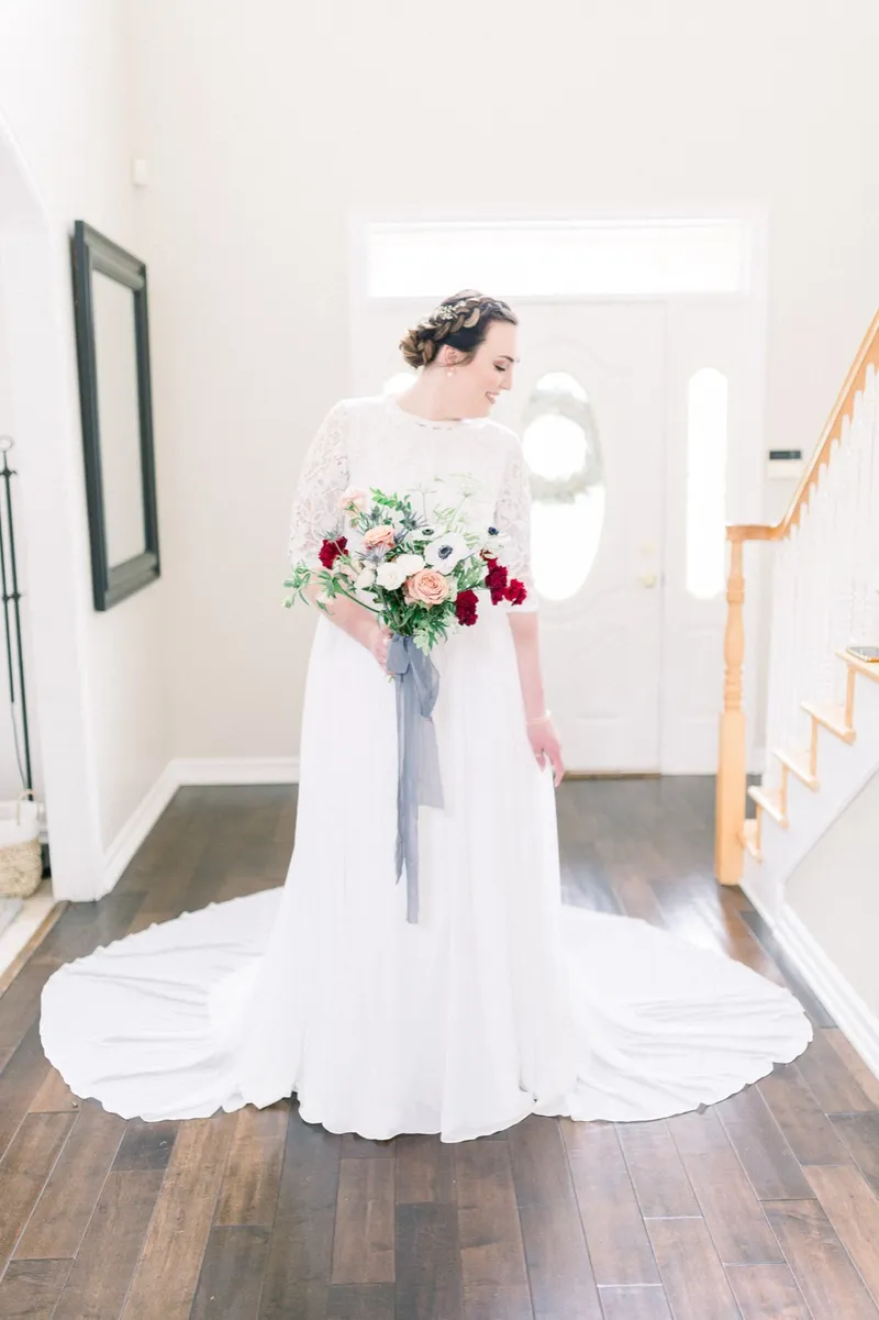 Bride in a lace sleeve gown standing by the staircase inside the house, holding a red and peach bouquet