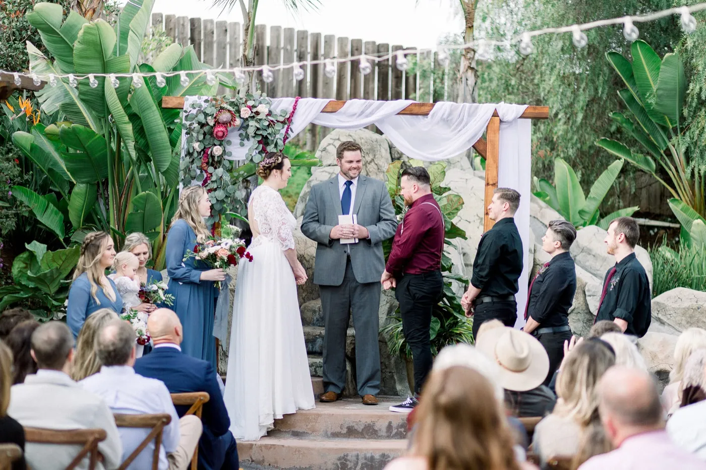 A couple stand with their officiant under a draped wooden arch in a backyard, guests seated in front of tropical plants