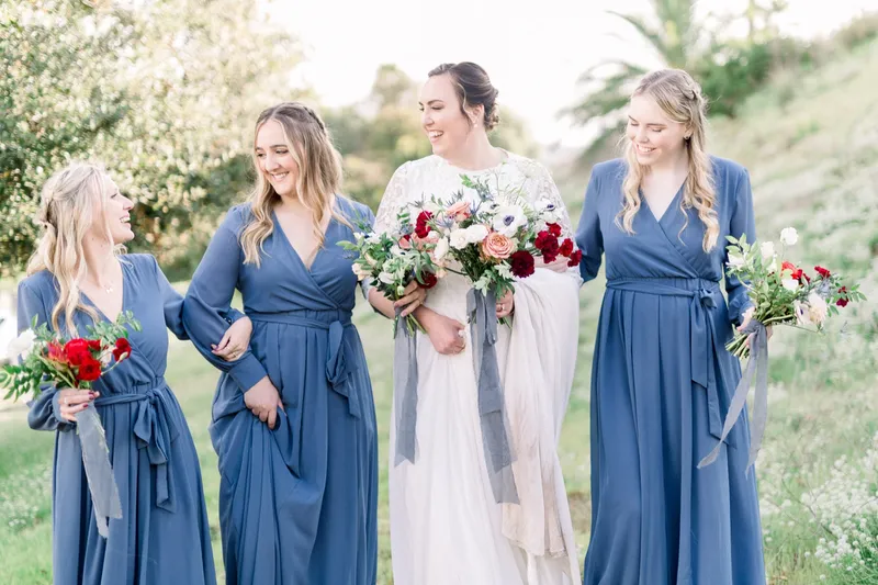 Bride with three bridesmaids in blue on a sunlit lawn, all holding red and peach bouquets