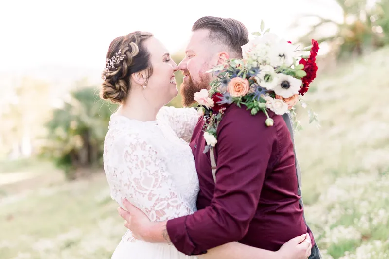Couple laughing close together with the bride's red and peach bouquet raised beside their faces