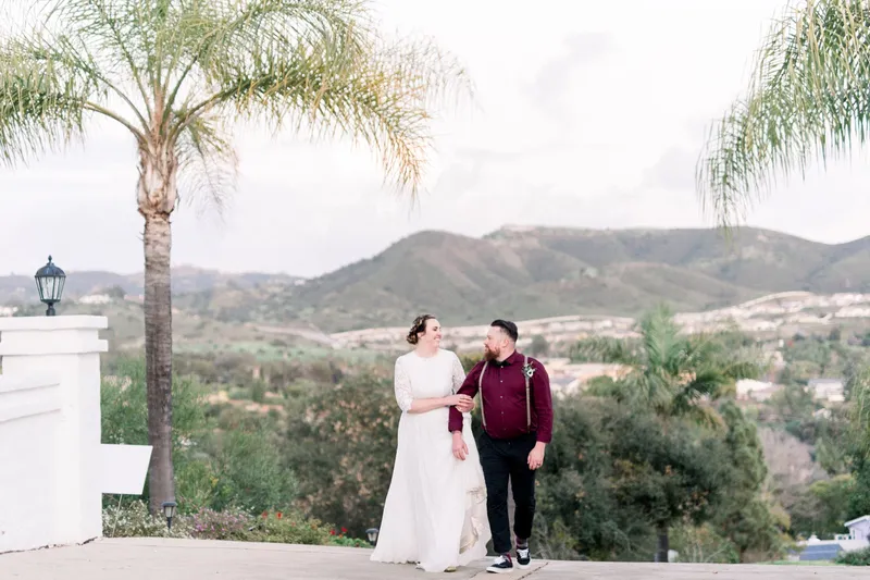 Couple walking hand in hand up the driveway past a row of palms, hills and a valley full of houses spread out behind them