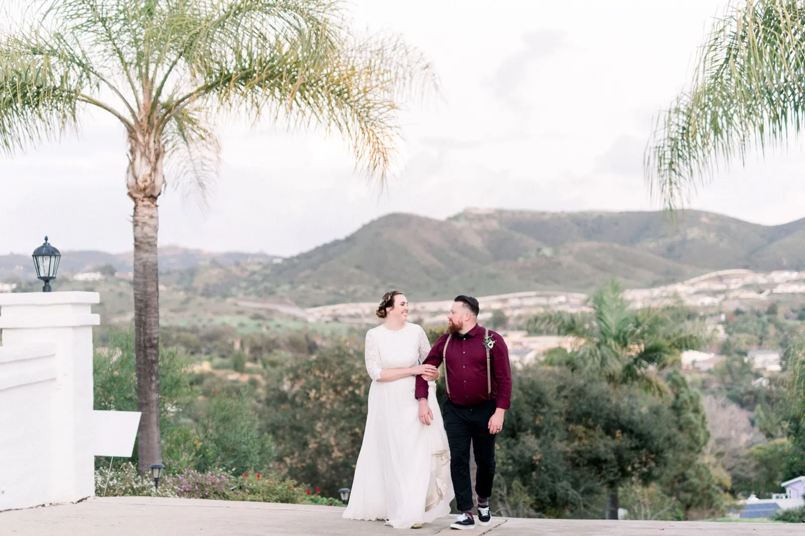 A couple walk hand in hand up a driveway lined with palms, hills and a valley of houses behind them