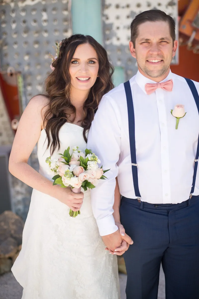 Bride holding a blush bouquet beside her groom in suspenders and a coral bow tie