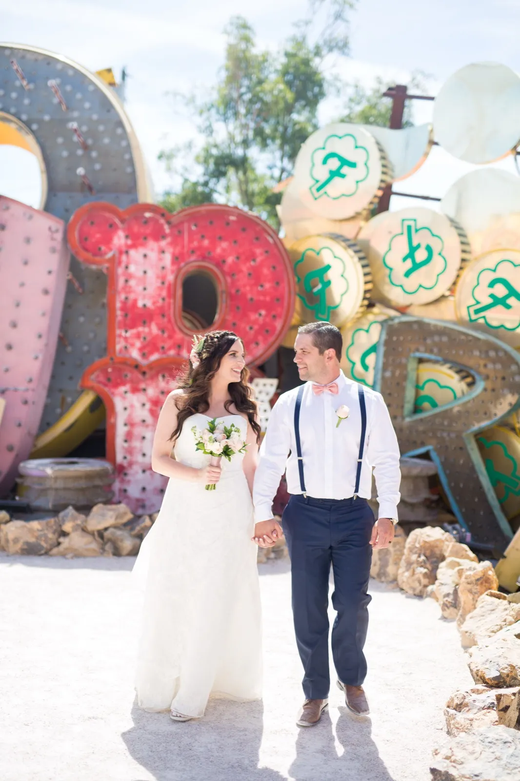 Bride and groom walking hand in hand up a gravel path between a giant red letter and stacked green and yellow signs