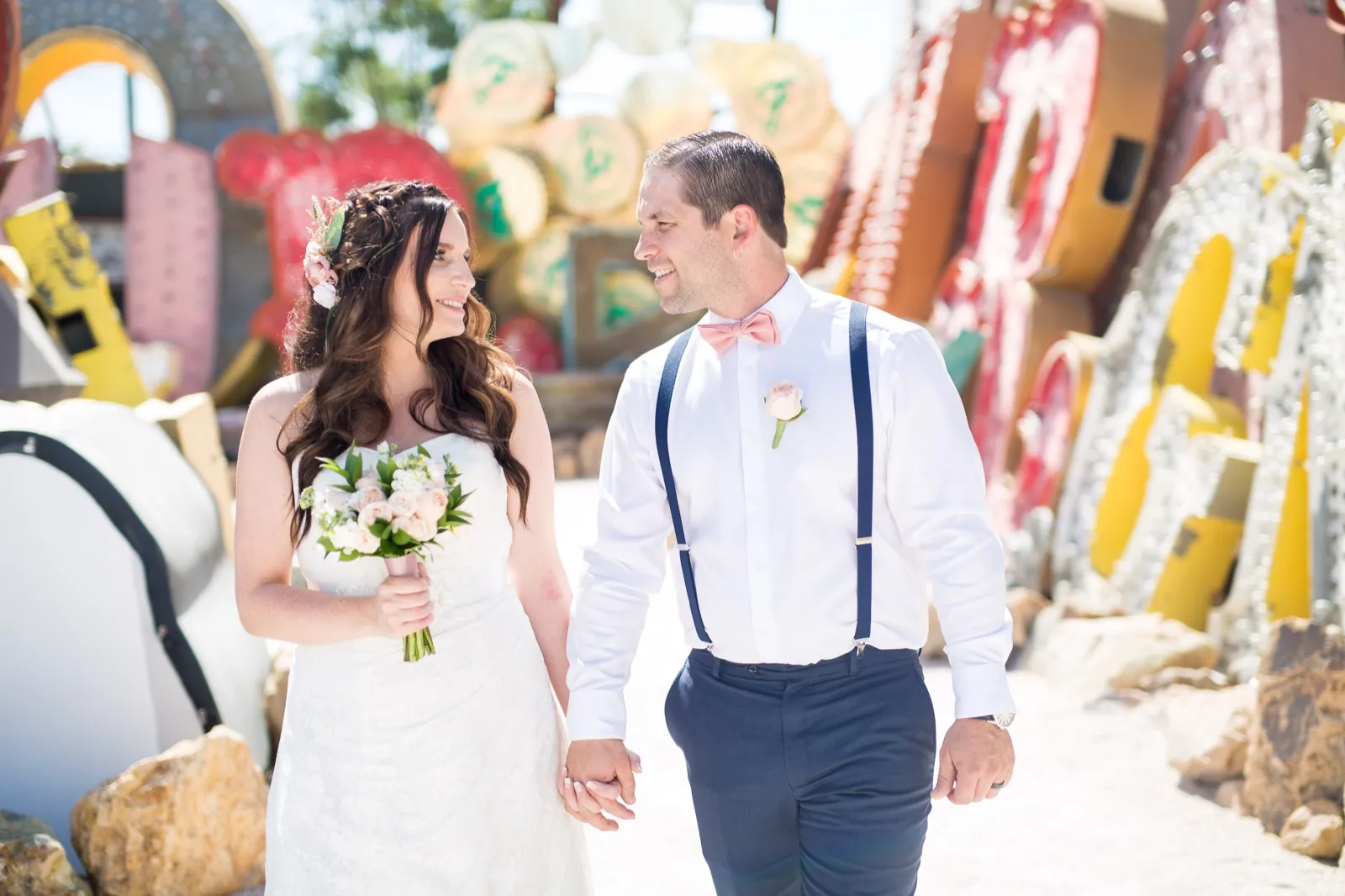 A bride with a bouquet and her groom in navy suspenders walk hand in hand past rows of old neon signs