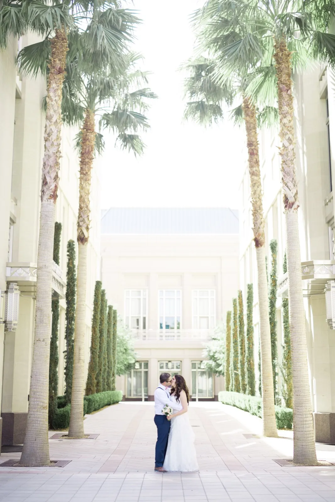 Couple kissing at the far end of a walkway lined with tall palms and cypress between the pale stone buildings