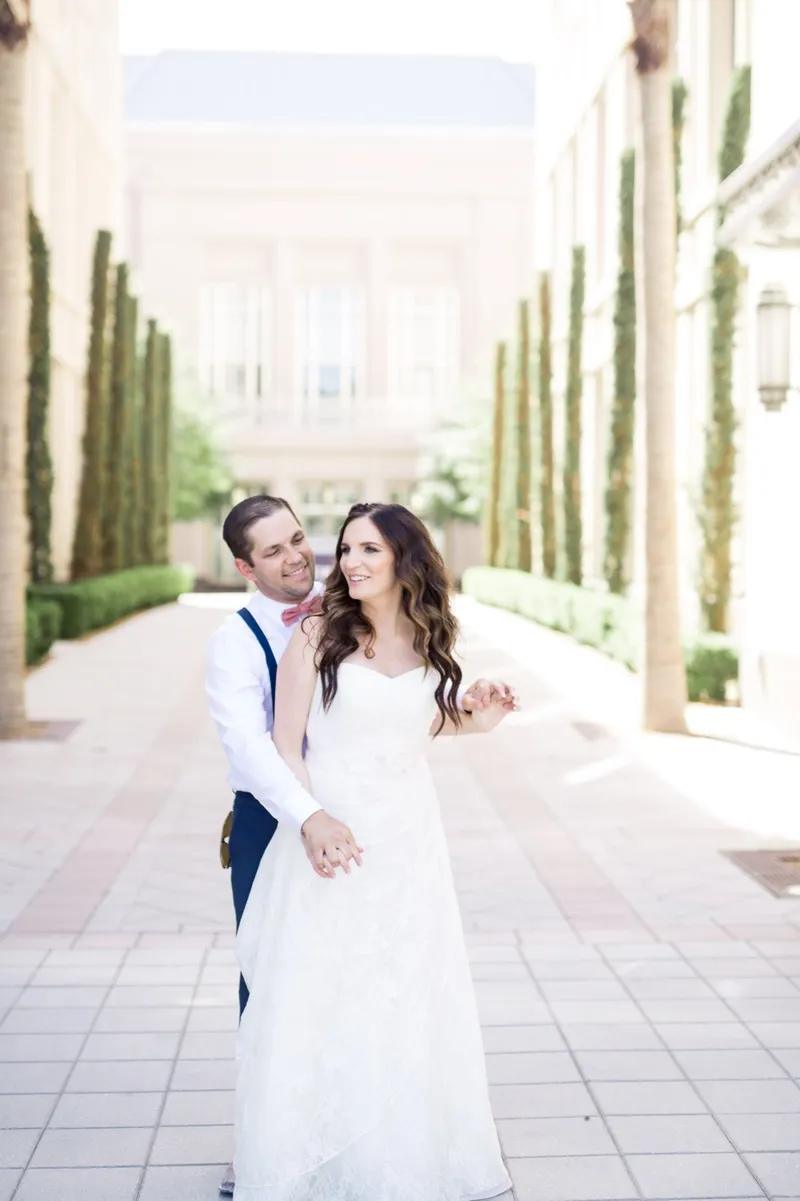 Bride in a lace gown and groom in white shirt and suspenders walking toward the camera down the cypress lined walk
