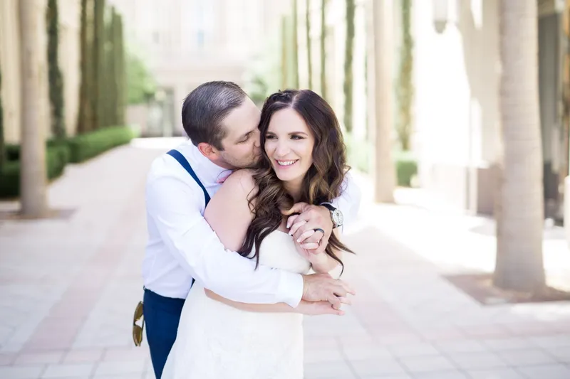 Groom kissing his fiancee on the cheek while she laughs with her arms wrapped around him, the pale stone plaza open behind them