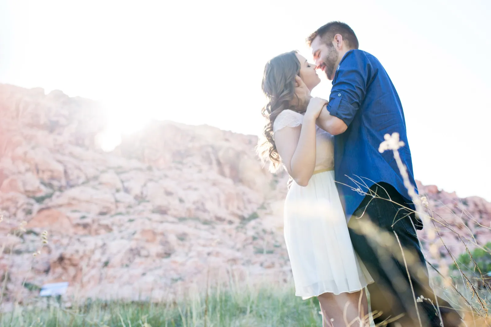 Couple about to kiss in tall grass as low sun flares across a pale red rock hillside behind them