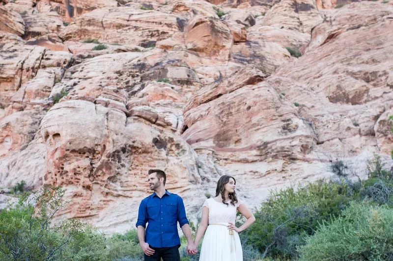 Couple standing apart and holding hands on a green slope below a tall red and cream rock face