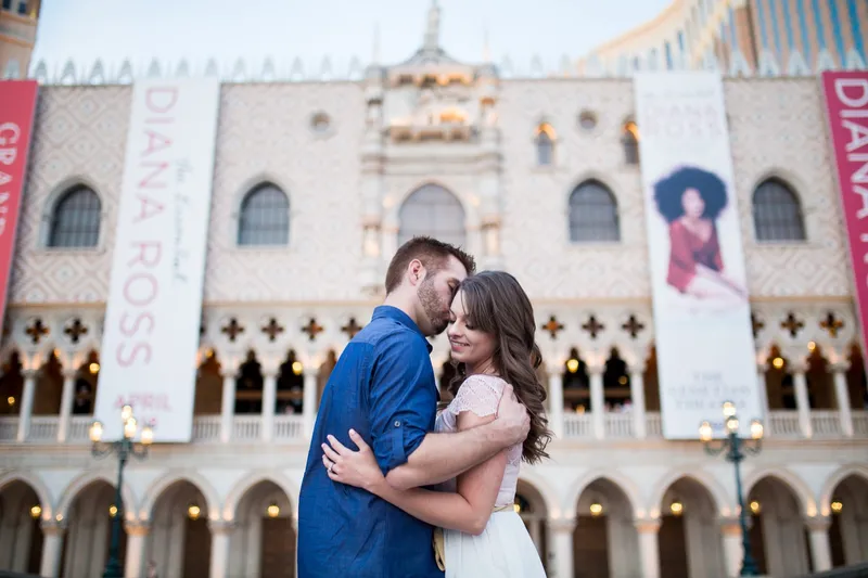 Couple holding each other at dusk with the lit Doge's Palace facade and its theater banners filling the frame behind them
