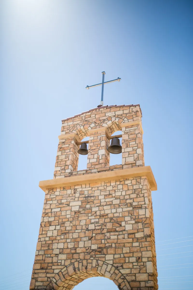 The freestanding stone bell tower with two bells hanging in its arches and a cross on top against a deep blue sky