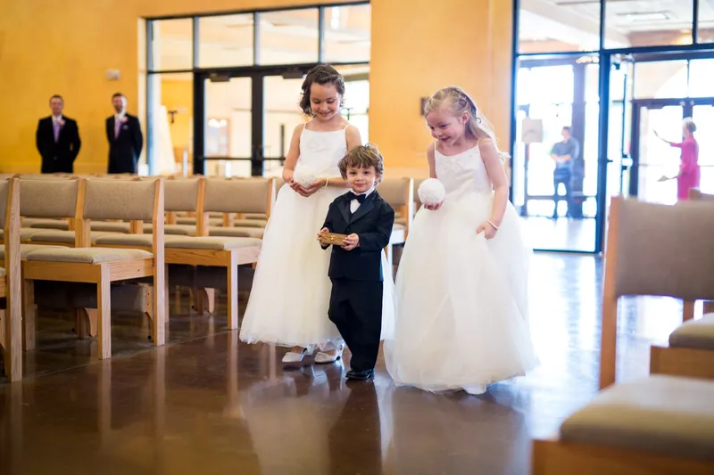 Two flower girls in ivory dresses and a small ring bearer in a black suit walking in together across the church floor